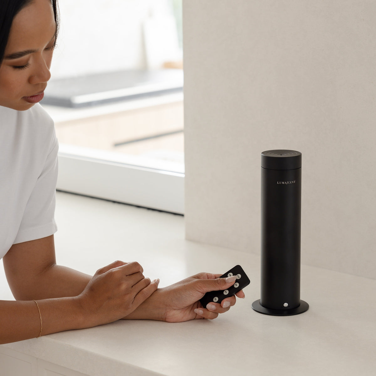 A woman in a white t-shirt sits at a white kitchen counter next to a black cylindrical smart speaker, holding a matching black remote control.