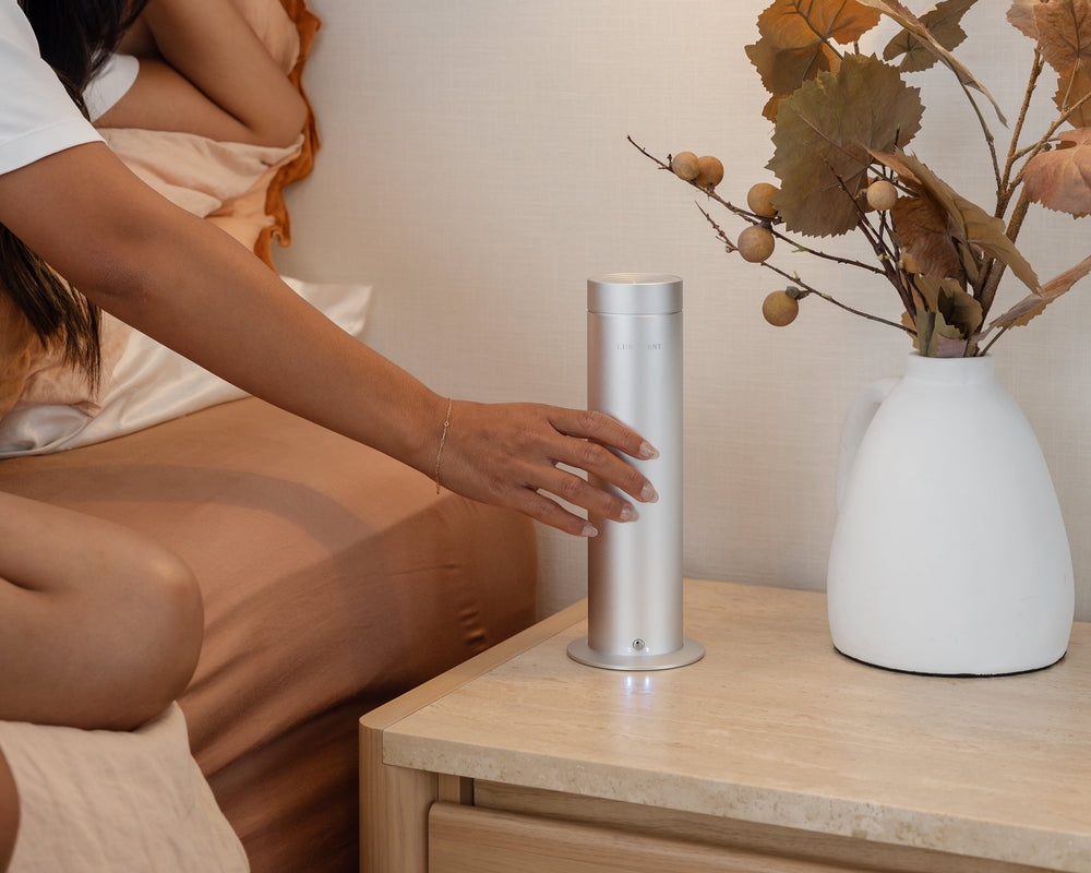 Woman sitting on a bed using a handheld device next to a nightstand with a white lamp and decorative plant.