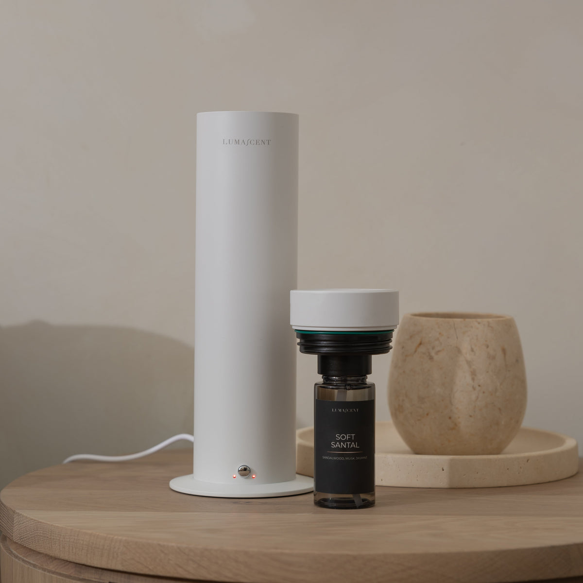 A white cylindrical smart water filter device standing on a round wooden table next to a small dark bottle, with a textured beige vase in the background.